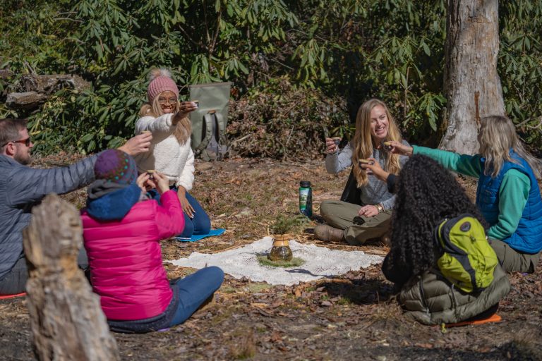 Five people sit in tea ceremony circle raising their tea cups in the air toasting at one another, displaying a unique wellness retreat near me experience.
