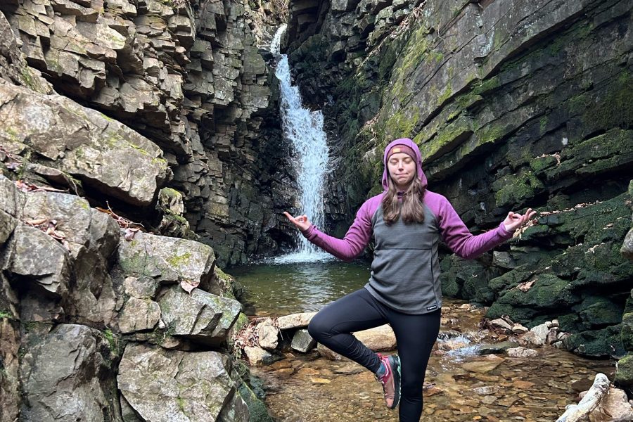 Women in purple and grey hoodie stand in tree pose in front of waterfall at one fo the best hikes in Smoky Mountains.