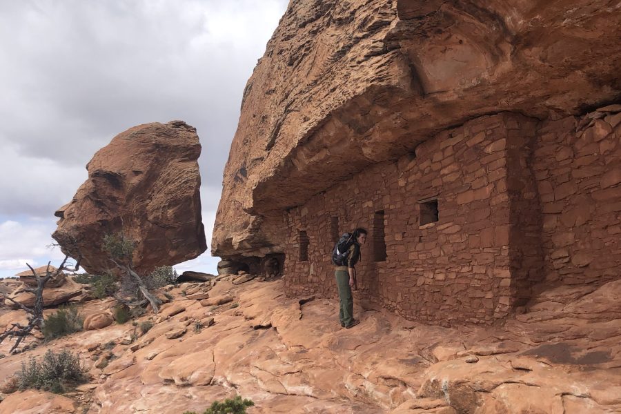 Backpacker in on a rock ledge while hiking Bears Ears National Monument of an ancient indigenous shelter built into a rock face on an overcast day.