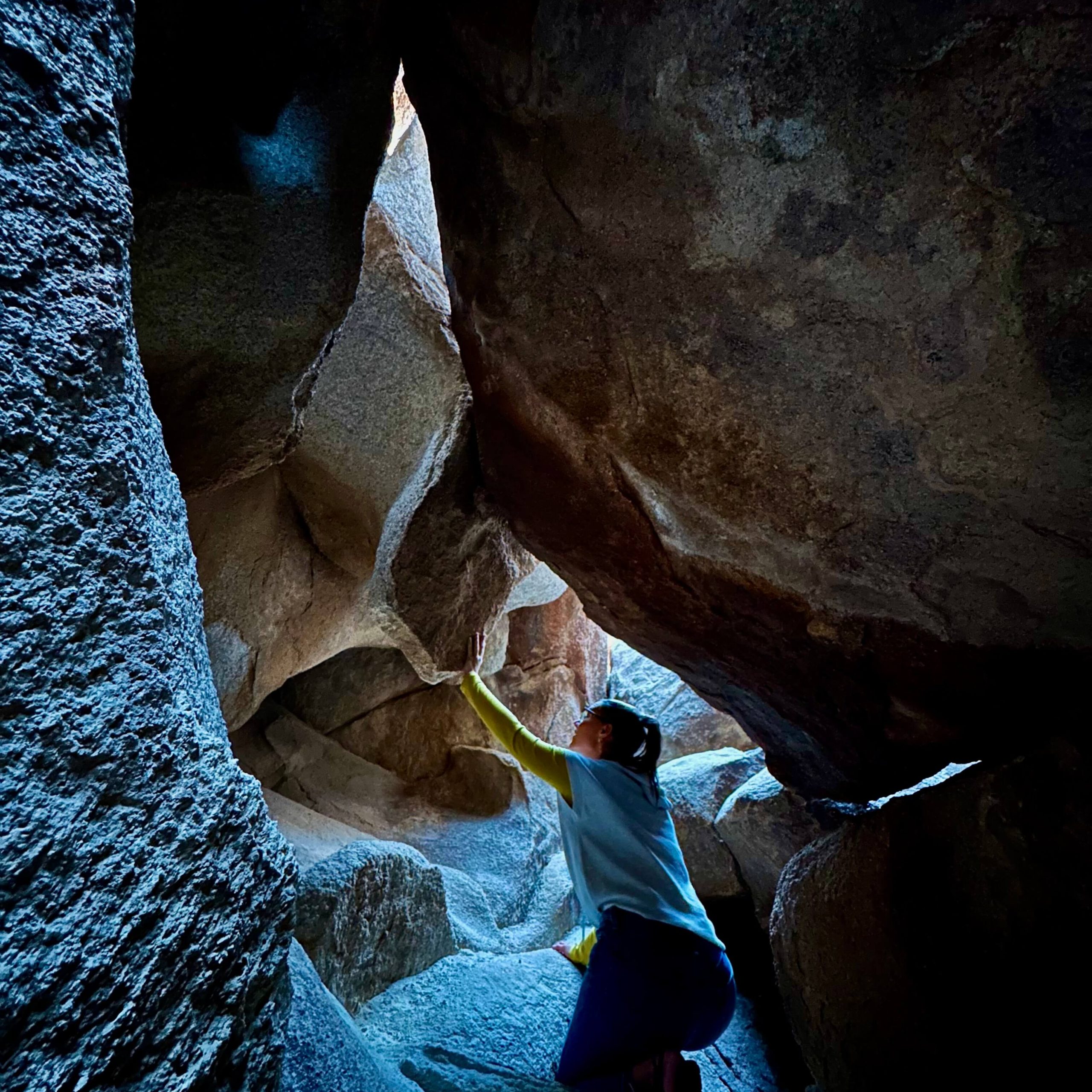 A women in white short sleeve shirt with yellow long shirt underneath crawls on her knees in a rock corridor. Her left arm is outreached on the ceiling of the corridor looking in awe.