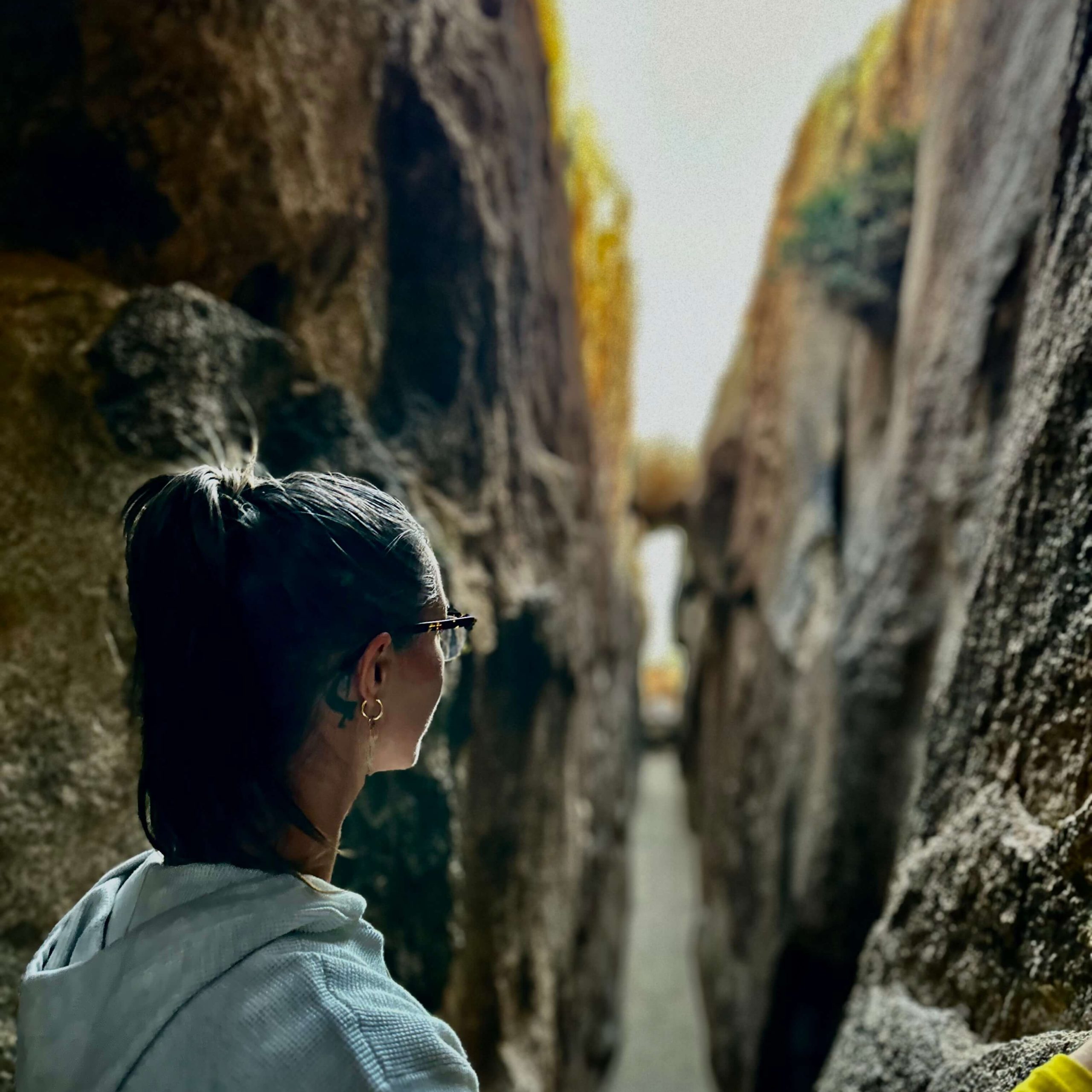 A view of the back of women's head as she gazes down bright rock corridor. She wears glasses, and grew hooded sweatshirt, and has her medium length brown hair in pony tail, showing a semi-circle black tattoo behind her right ear.