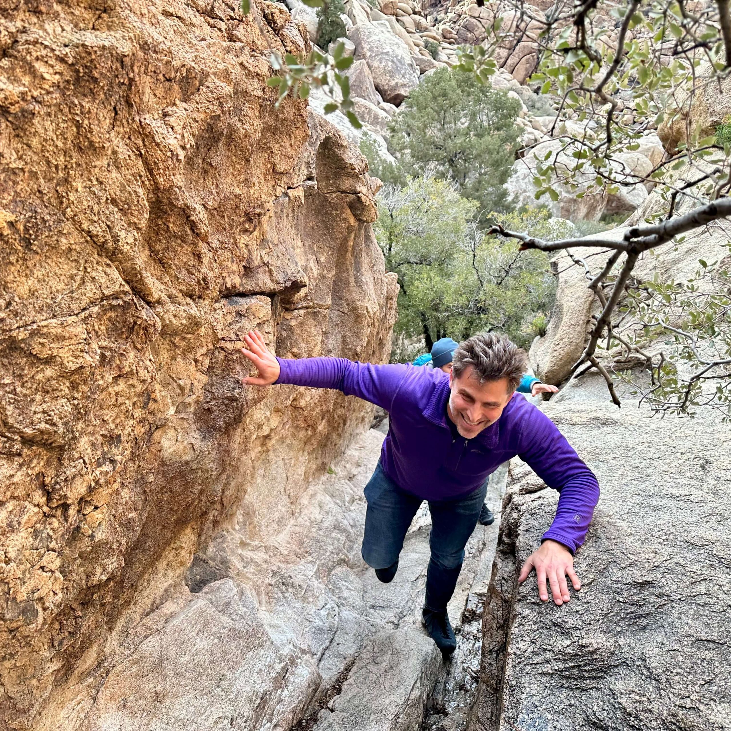Man in purple long sleeve and jeans with brown hair climbs toward camera on a rock corridors entrance.