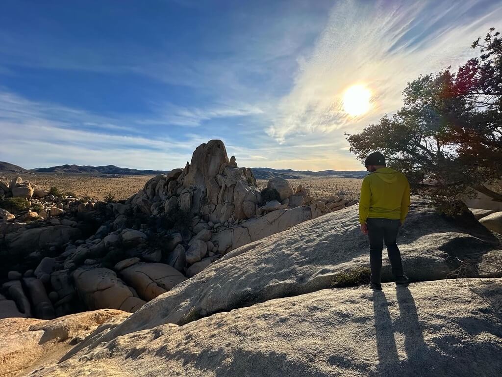 A person in yellow long sleeve and grey pants with right leg cocked out, stands on a rock face looking down at the corridor below during more sunrise on southern California hike.