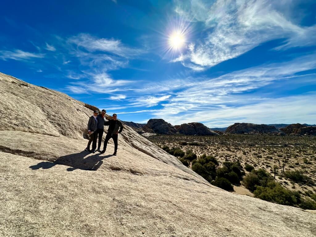 Three people in sun protective clothing stand on a rock face slanting at a downward angle from the left to right. There is a desert scape to the right with a sunny blue-skied day.