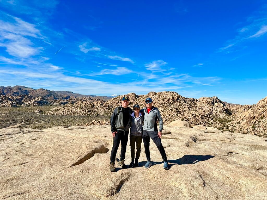 A group of three people pose on a rock with desert rock outcroppings in the distance. There is a man on each side with a women in the middle, all of which are wearing long-sleeve and pants with sun protection.