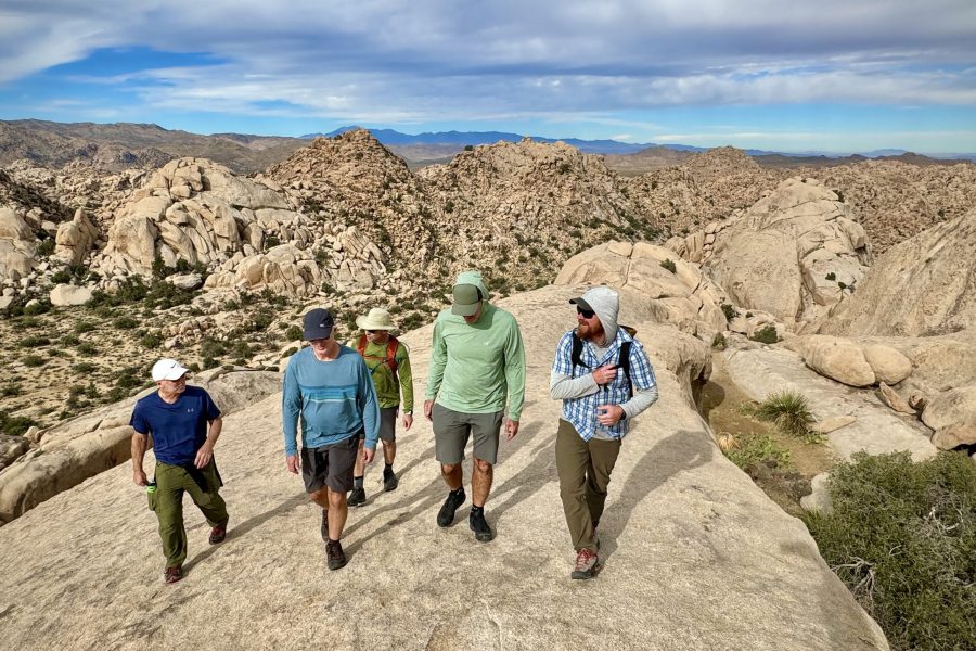 A Joshua Tree hikes guide is walking across a large tan rock with a group of four clients teaching them about the park. The day is partly cloudy with blue skies peaking through the clouds and a long view of the desert in the background.