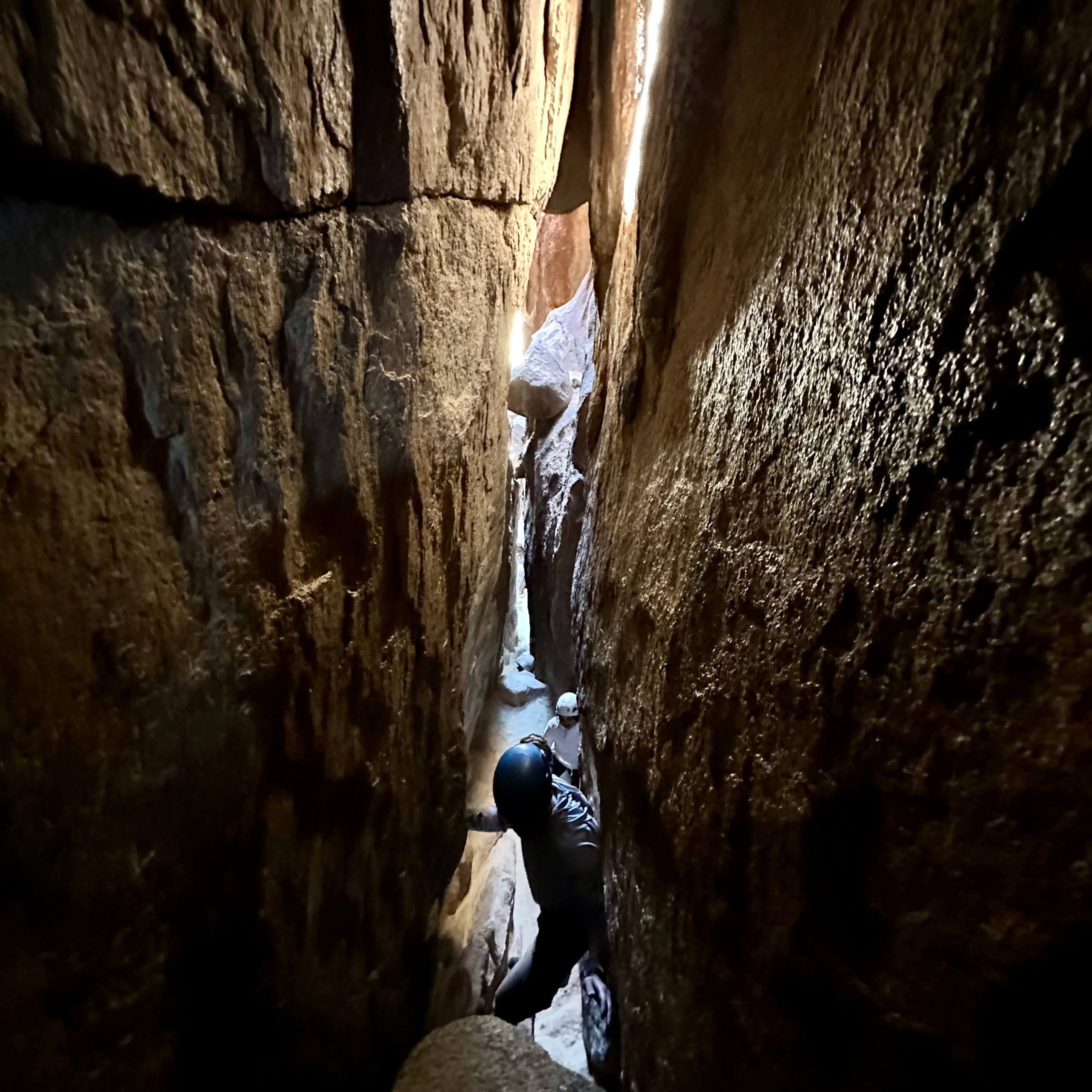 A dark view looking down into a dimly lit rock corridor of two people shifting sideways through the passage.