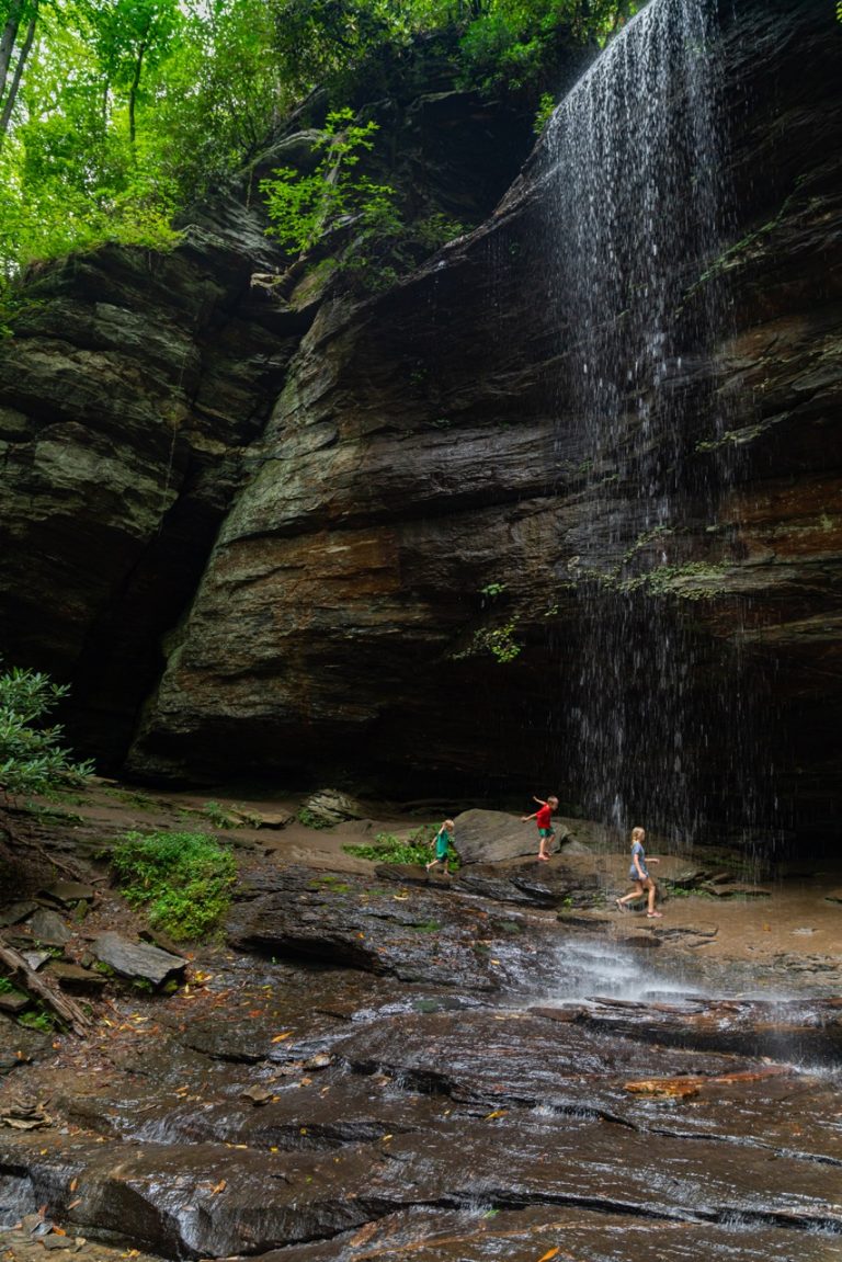 Children playing under the rock ledge of a waterfall, which is one of the best hikes near Asheville when hiking the blue ridge mountains.