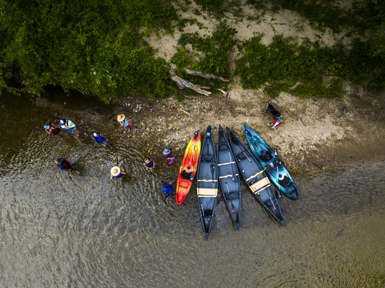 Aerial view of five kayaks during an French Broad River kayak tour along the river shore with a group of people engaging in outdoor activities and enjoying a summer day in the water.