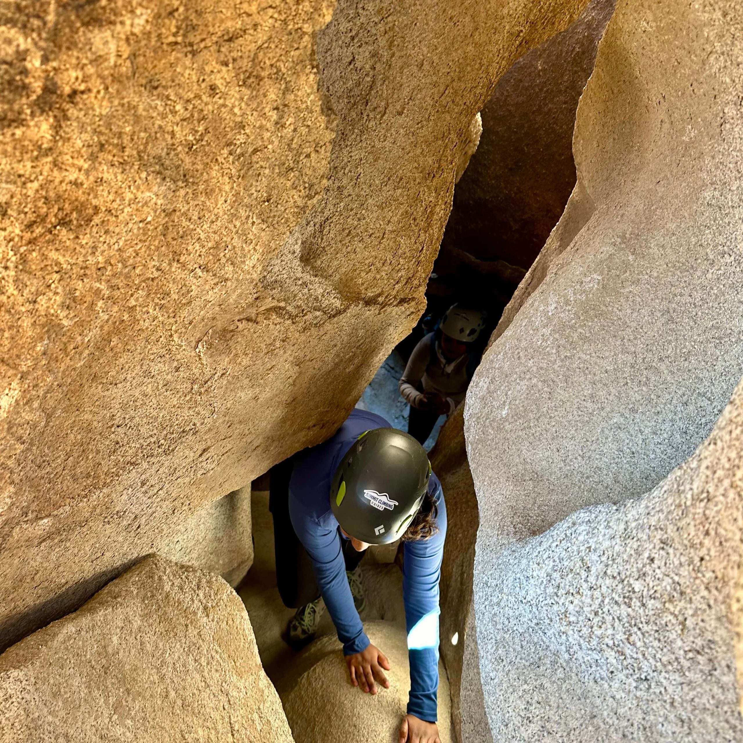 A person with blue long sleeve shirt and black helmet crawls on all fours through tight corridor toward the exit while the rest of the group follows.