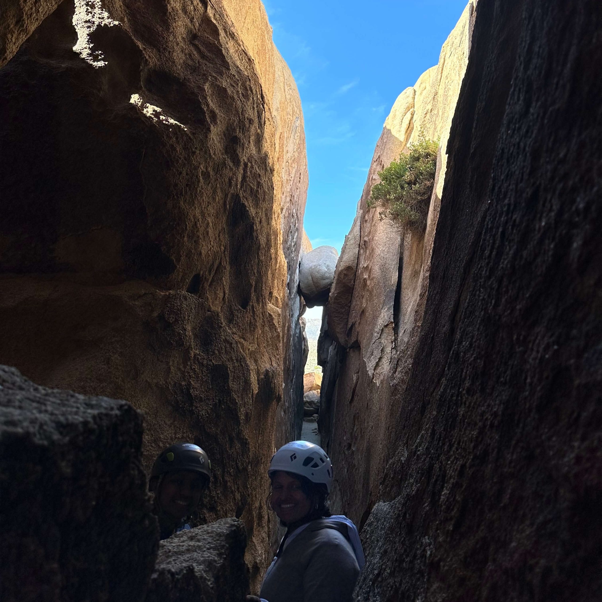Two people with helmets stand in body-width rock corridor posing for picture during adventure cave tour with blue sky beyond the labyrinth.