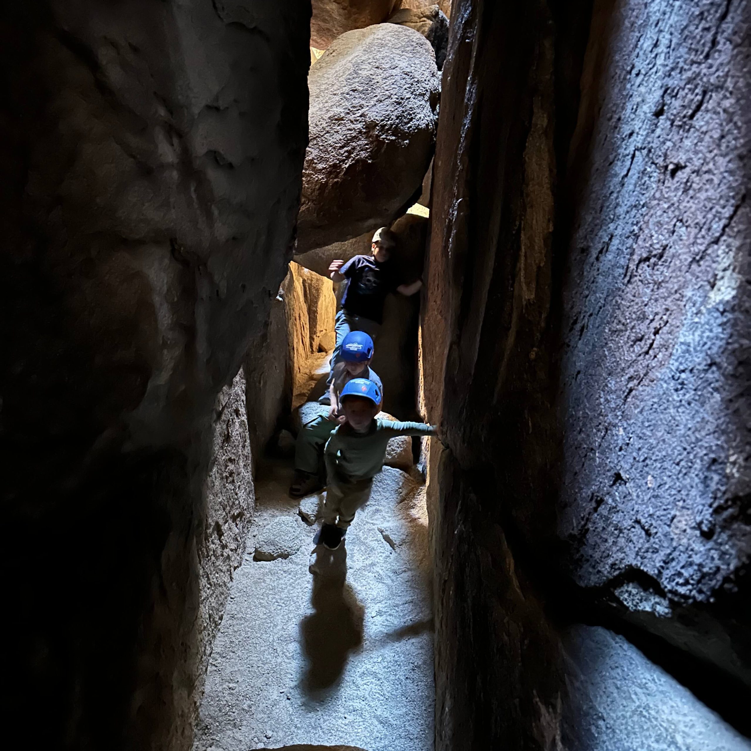 A group of children with safety helmets on walk toward the camera in a sun beam shining down in a dark cave corridor.