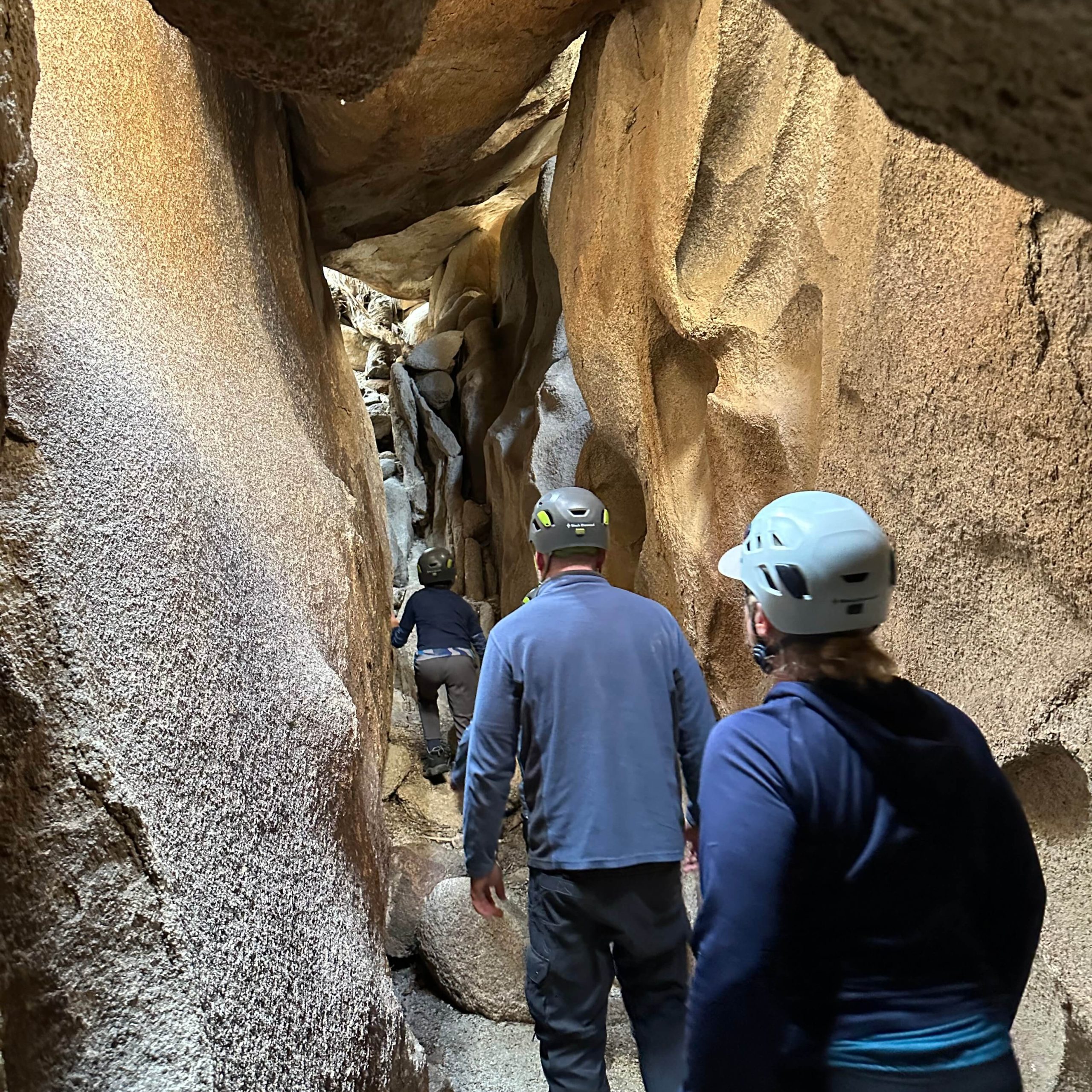 A family of three, one women, man, and small child in helmets wander between small corridor tour to cave beyond. The corridor is brightly light with light brown rock on both sides.