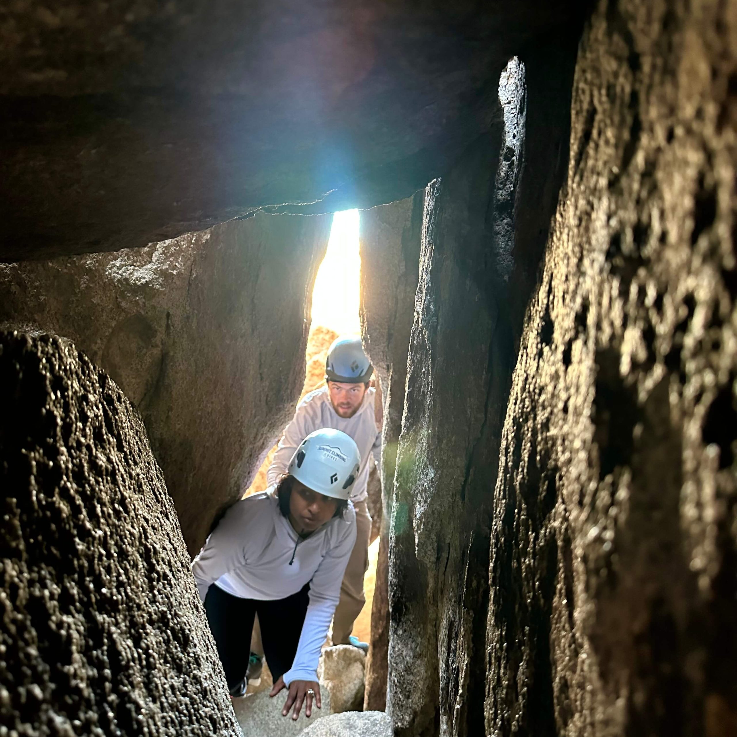 Two explorers with helmets on crawl through narrow opening in rock corridor.
