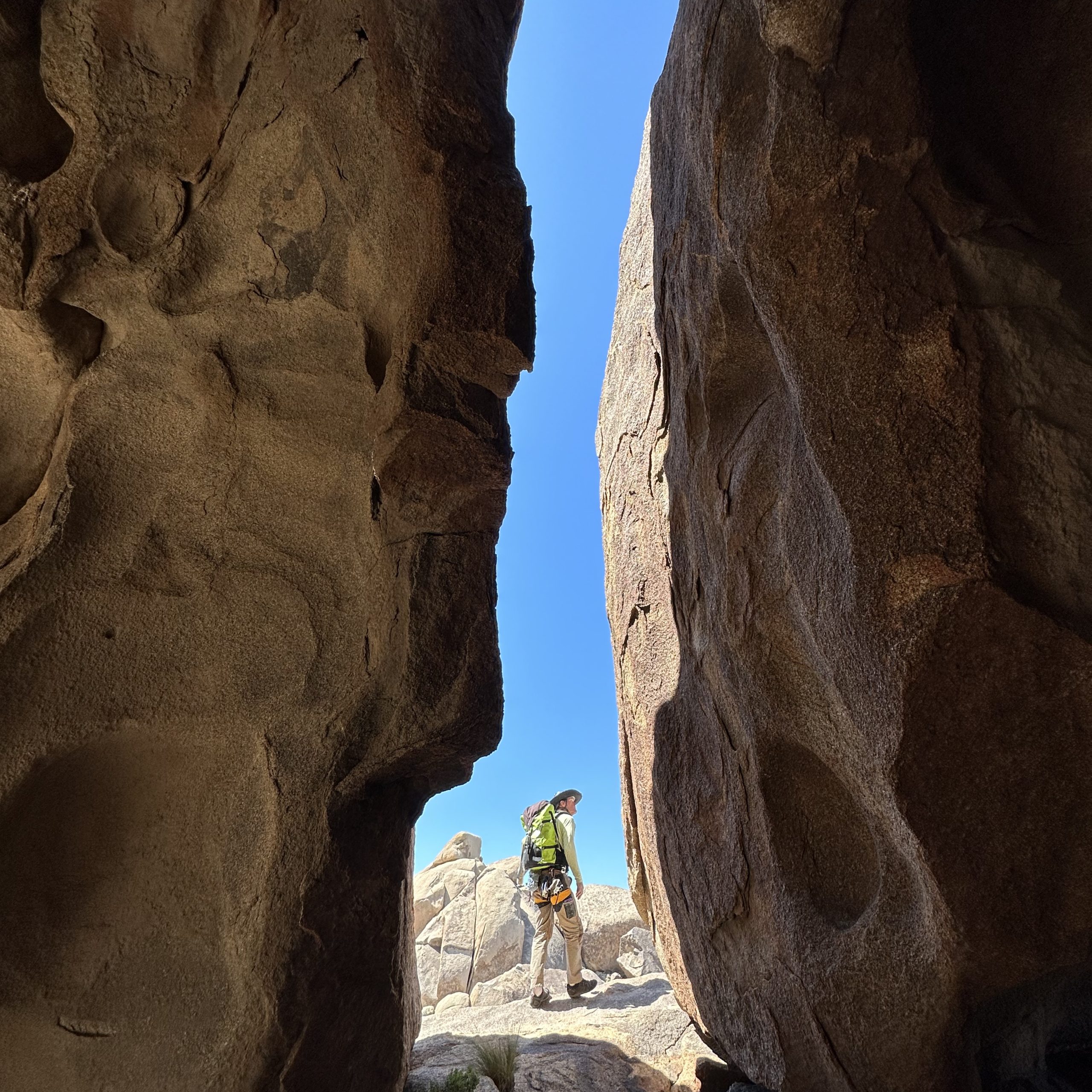 A hiker with green backpack, pants, and hat is seen through a vertical opening leading away from a Joshua Tree cave between two boulders with blue skies beyond.