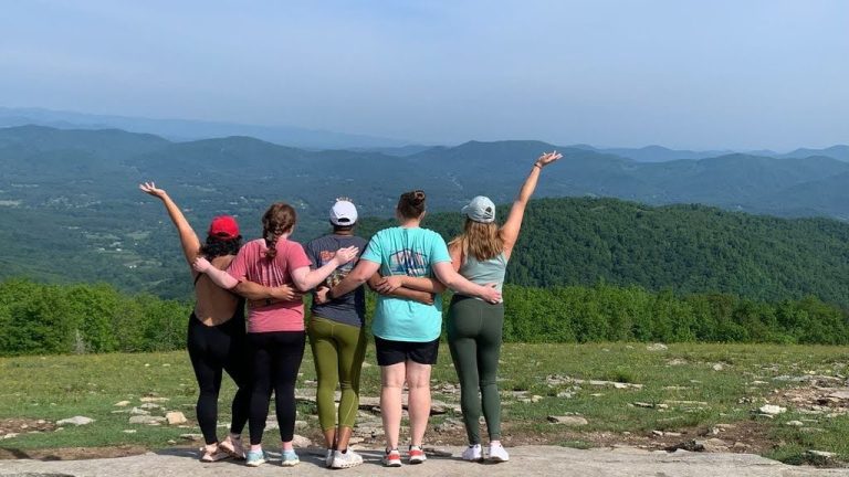 Five women in yoga apparel after yoga for hikers class stand on a rocky hill overlooking a view of Blue Ridge Mountains on sunny day.