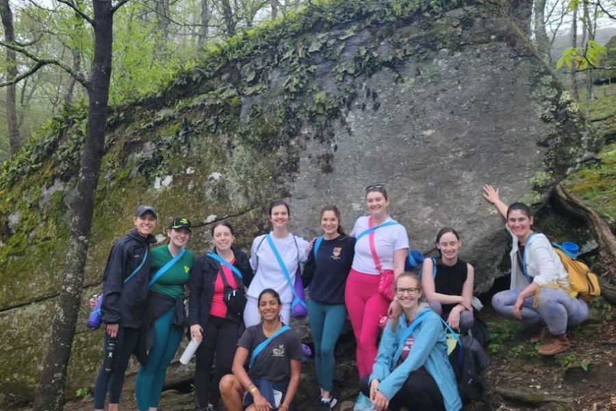 Ten people stand in yoga clothes with yoga mat slung around their back at the base of large mossy boulder after nature yoga as they smile for the camera.