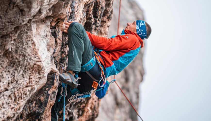 Man in a blue bandana and red-blue jacket rock climbing a rugged face, showcasing a thrilling outdoor activity