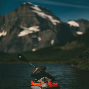 person with hat in red and grey kayaking toward snowy mountains